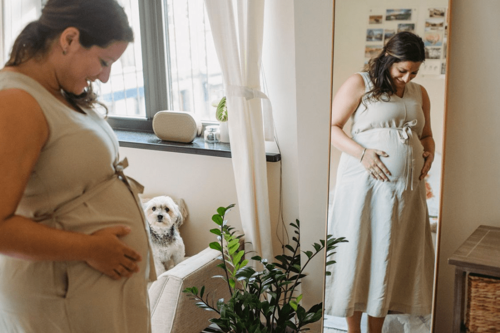 A pregnant woman in a beige dress gently cradles her belly while looking at her reflection in a mirror, with a small dog sitting on a couch nearby.